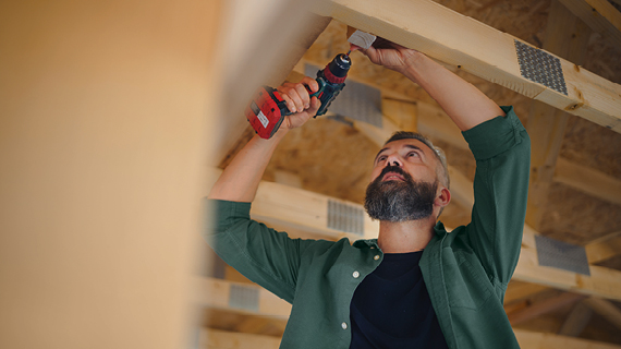 Person arbeitet mit einer Bohrmaschine an einem Holzbalken in einem Gebäude.“ class=