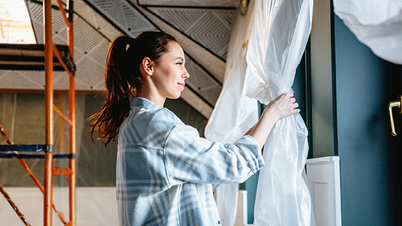 Person in kariertem Hemd zieht eine Plastikfolie von einer Wand in einem Raum mit Baugerüst und schräger Decke.