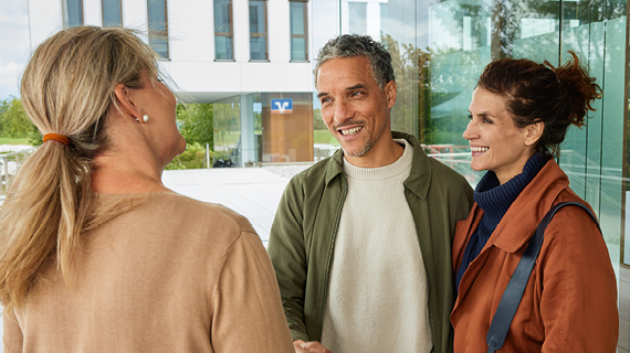 Drei Personen stehen vor einem modernen Gebäude und unterhalten sich; eine Person schüttelt einer anderen die Hand.