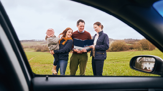 Drei Erwachsene und ein Kind stehen auf einem Feld und betrachten gemeinsam ein Dokument; das Bild ist durch das geöffnete Fenster eines Autos aufgenommen.“